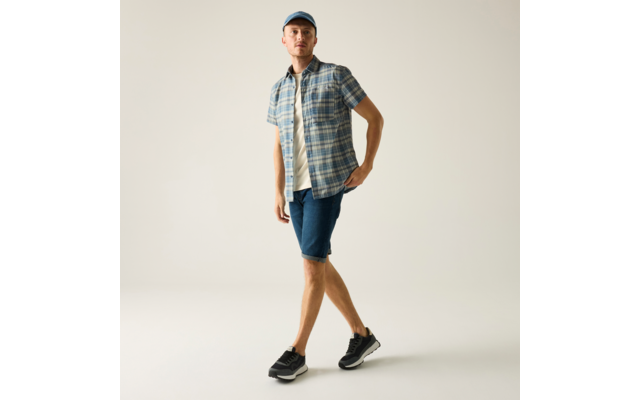 Mann mit blauer Kappe, kariertem Kurzarmhemd über weißem T-Shirt, Jeans-Shorts und schwarzen Sneakers vor hellem Hintergrund