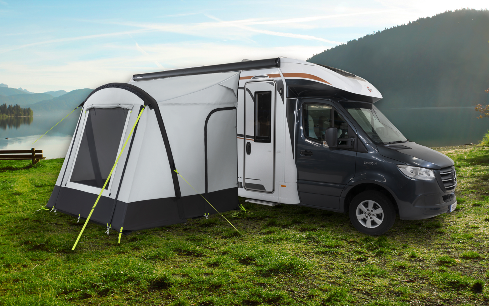 Camper van with attached tent on a green meadow near a lake and mountains in the background