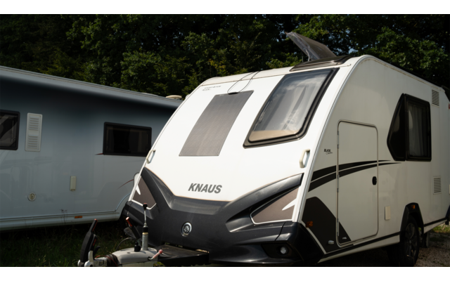 Caravana blanca con logo KNAUS negro, ventana frontal, enganche de remolque y ventana lateral; al fondo otra caravana y árboles