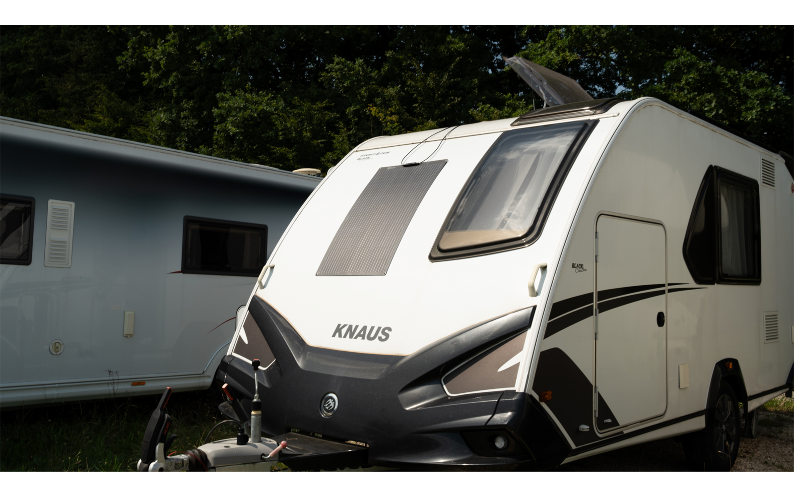 Caravana blanca con logo KNAUS negro, ventana frontal, enganche de remolque y ventana lateral; al fondo otra caravana y árboles