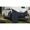 Parte frontal de una caravana blanca con la palabra "KNAUS", un panel solar apoyado delante, hierba y grava en primer plano, enganche de remolque visible