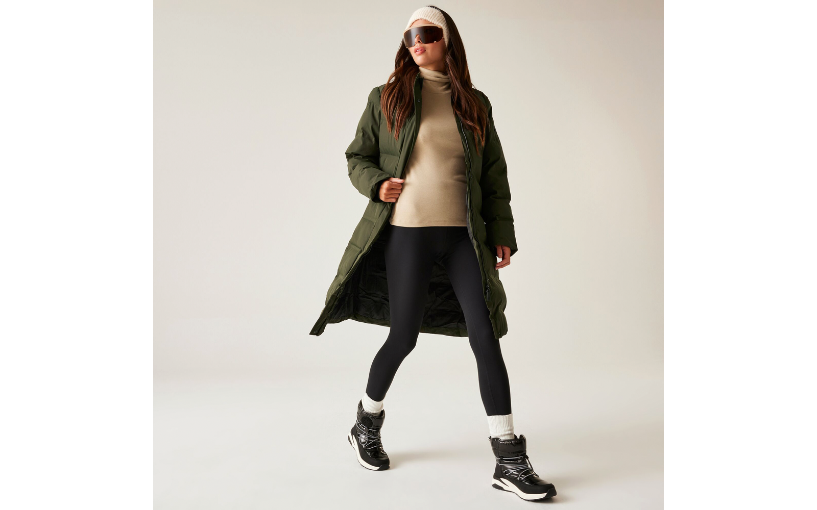 Frau mit Sonnenbrille, langer grüner Jacke, beigem Pullover, schwarzer Leggings und schwarzen Stiefeln beim Gehen