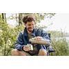 A person with short curly hair sits on a folding chair at the water's edge, pouring from a stainless steel thermos into a mug
