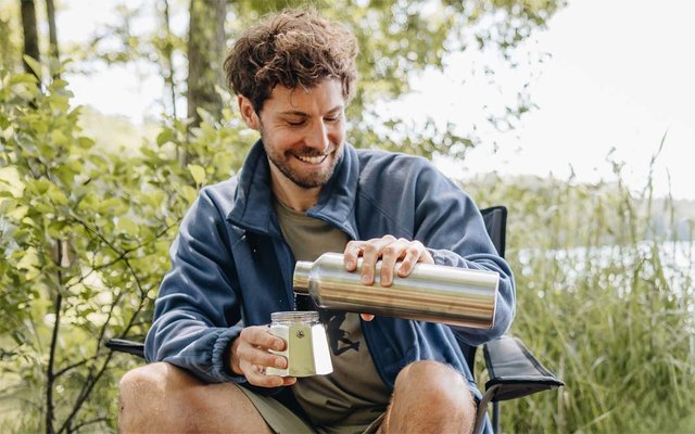 A person with short curly hair sits on a folding chair at the water's edge, pouring from a stainless steel thermos into a mug