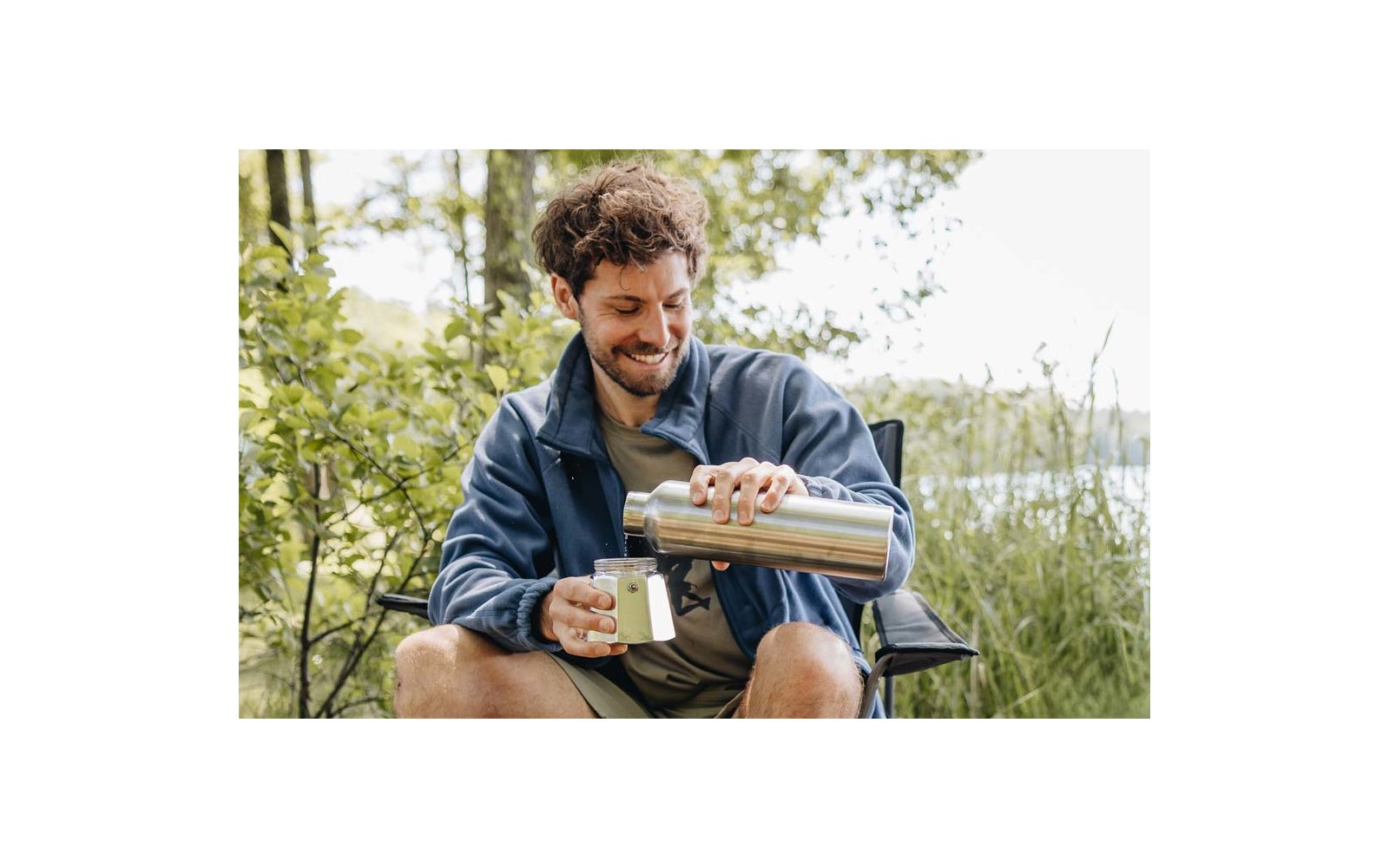 A person with short curly hair sits on a folding chair at the water's edge, pouring from a stainless steel thermos into a mug