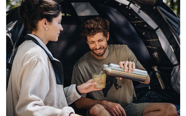 Two people sit in front of an open tent; one person pours liquid from a stainless-steel flask into a glass held by the other person