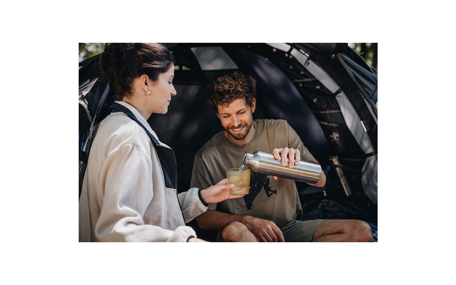Two people sit in front of an open tent; one person pours liquid from a stainless-steel flask into a glass held by the other person