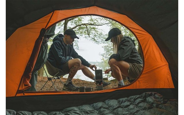 Blick aus einem orangen Zelt auf zwei Personen in Outdoor-Kleidung, die auf Campingstühlen neben einem kleinen Kocher sitzen; Bäume und Wasser im Hintergrund; Schlafsack im Zelt sichtbar