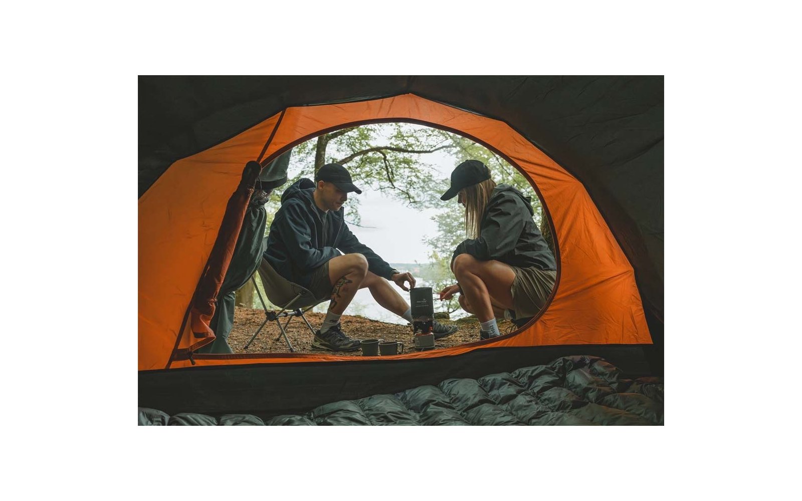 Blick aus einem orangen Zelt auf zwei Personen in Outdoor-Kleidung, die auf Campingstühlen neben einem kleinen Kocher sitzen; Bäume und Wasser im Hintergrund; Schlafsack im Zelt sichtbar