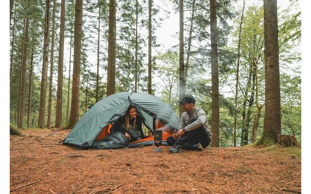 Grünes Kuppelzelt in einem Nadelwald, zwei Personen – eine im Zelteingang, eine sitzt davor, kleiner Campingkocher mit Kessel und aufsteigendem Dampf, Waldboden mit Kiefernnadeln
