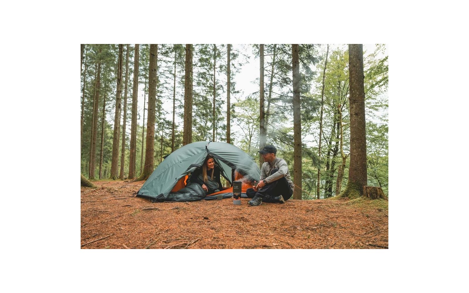 Grünes Kuppelzelt in einem Nadelwald, zwei Personen – eine im Zelteingang, eine sitzt davor, kleiner Campingkocher mit Kessel und aufsteigendem Dampf, Waldboden mit Kiefernnadeln