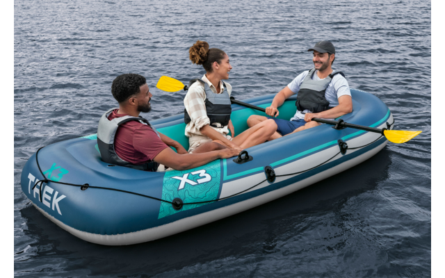 Drei Personen sitzen in einem blauen Schlauchboot auf offenem Wasser; zwei gelbe Paddel sichtbar; alle tragen Schwimmwesten