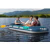 Drei Personen in einem aufblasbaren Boot mit der Aufschrift "TREK" auf einem See, ein gelbes Paddel, baumbewachsenes Ufer und Berge im Hintergrund