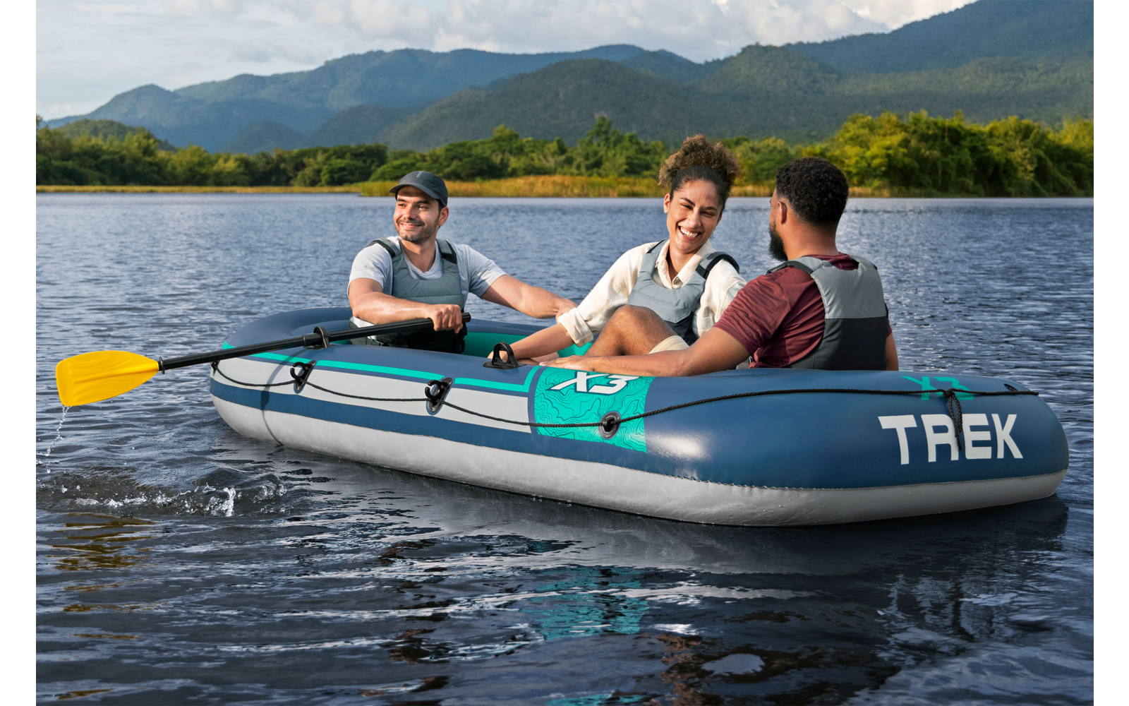 Drei Personen in einem aufblasbaren Boot mit der Aufschrift "TREK" auf einem See, ein gelbes Paddel, baumbewachsenes Ufer und Berge im Hintergrund
