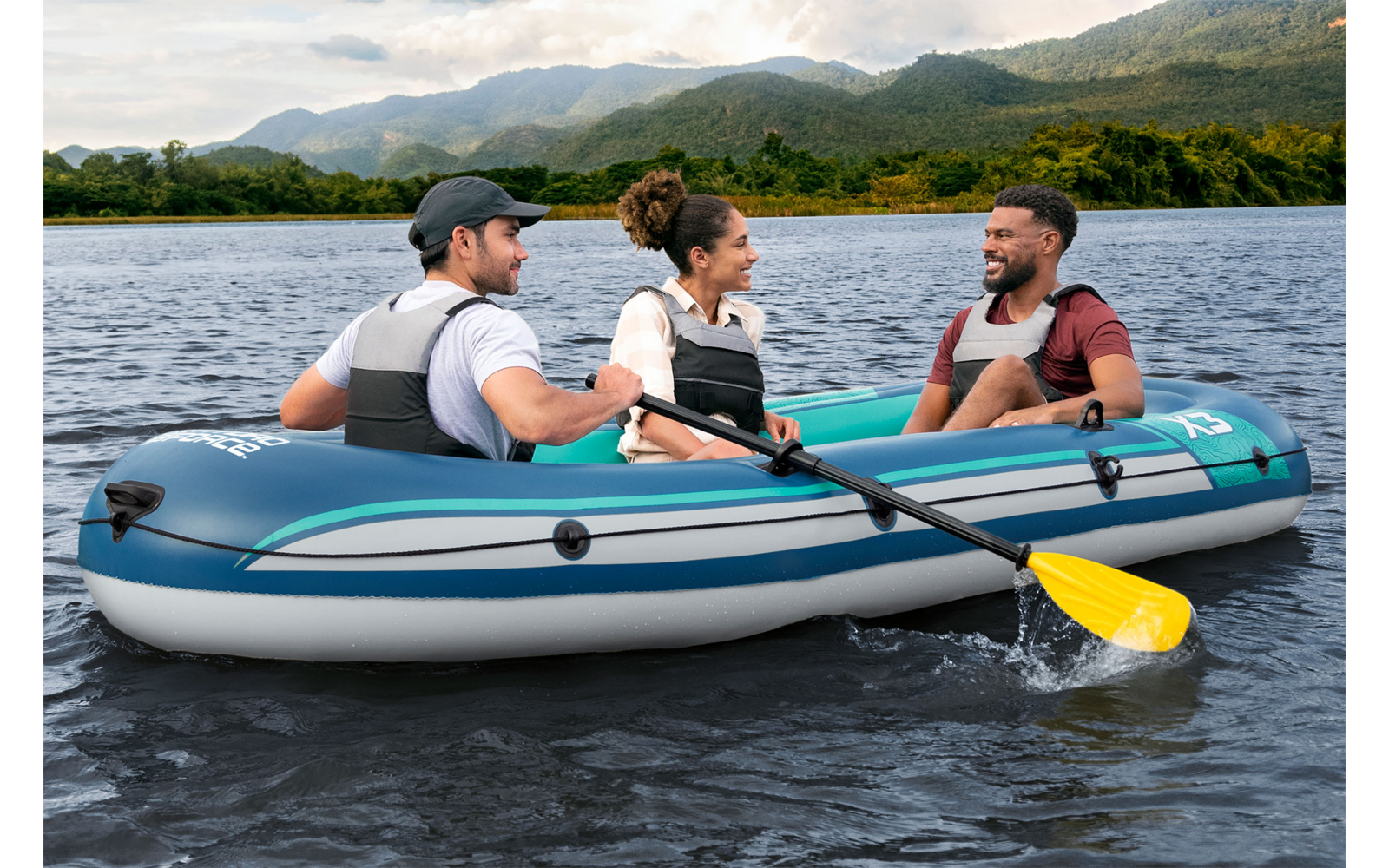 Drei Personen sitzen in einem blauen Schlauchboot auf dem Wasser; zwei halten Paddel, ein gelbes Paddel berührt das Wasser; alle tragen Schwimmwesten; bewaldetes Ufer und Berge im Hintergrund; wolkiger Himmel