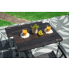 Table pliante rectangulaire foncée sur une nappe de pique-nique à motifs, deux verres avec liquide orange sur des dessous blancs, petit pot de plante et une grappe de raisins verts, deux accoudoirs de chaise, pelouse et chemin en arrière-plan