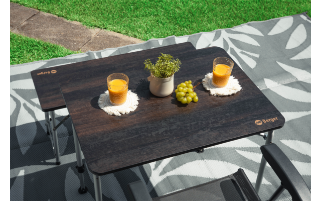 Table pliante rectangulaire foncée sur une nappe de pique-nique à motifs, deux verres avec liquide orange sur des dessous blancs, petit pot de plante et une grappe de raisins verts, deux accoudoirs de chaise, pelouse et chemin en arrière-plan