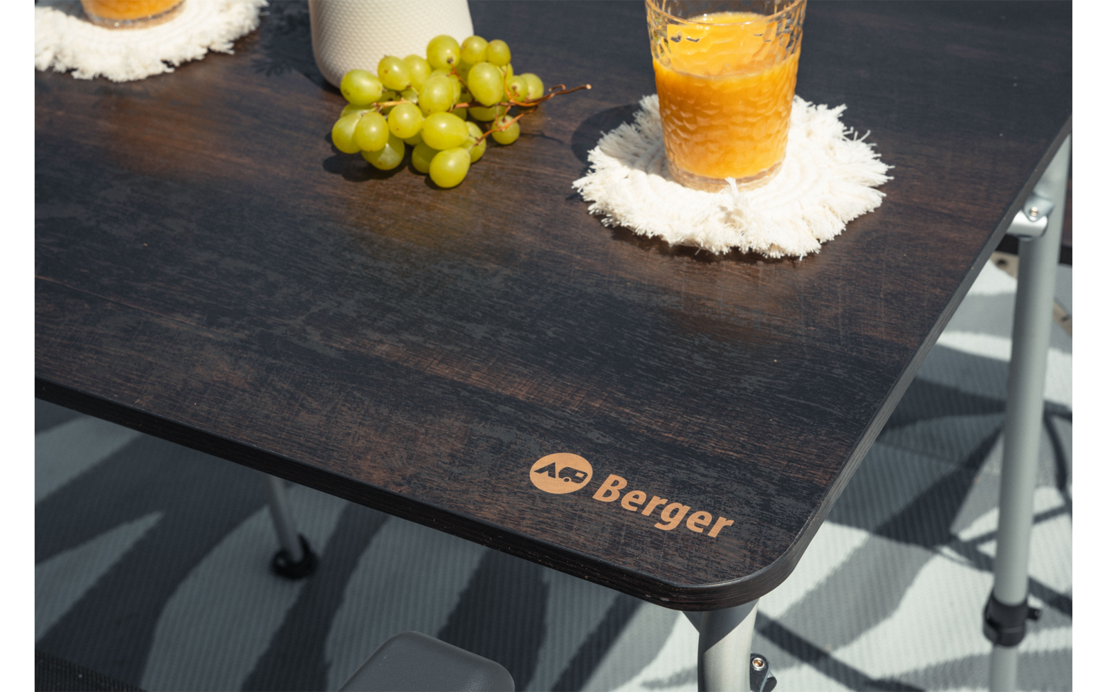 Plateau de table foncé avec raisins verts, verre de jus d'orange sur un dessous de verre blanc à franges, logo 'Berger' à l'angle, pieds métalliques et sol rayé visibles