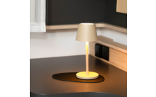 Beige Tischlampe mit eingeschaltetem Licht auf rundem Untersetzer auf schwarzem Schreibtisch, neben Holzkante und Steckdose; links Vorhang sichtbar