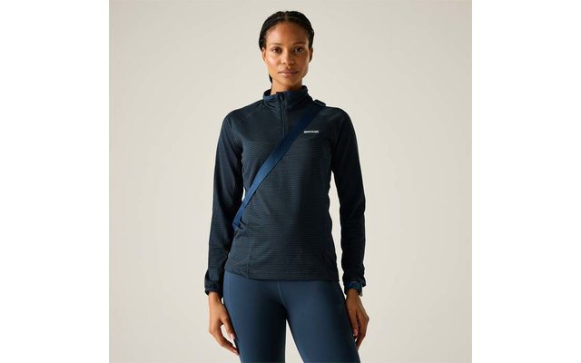 Personne debout de face, porte une veste zippée sombre à manches longues, leggings ajustés, bandoulière diagonale, cheveux nattés, fond clair
