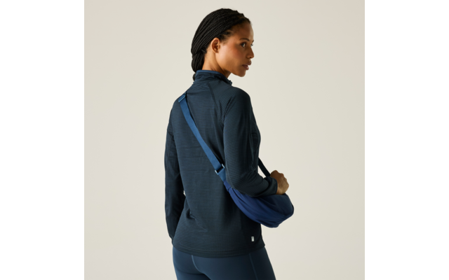 Femme de dos, légèrement tournée vers la gauche, portant un haut sombre à manches longues, des leggings bleu foncé et un sac bandoulière bleu en travers du dos, cheveux tressés, fond clair uni