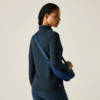 Femme de dos, légèrement tournée vers la gauche, portant un haut sombre à manches longues, des leggings bleu foncé et un sac bandoulière bleu en travers du dos, cheveux tressés, fond clair uni