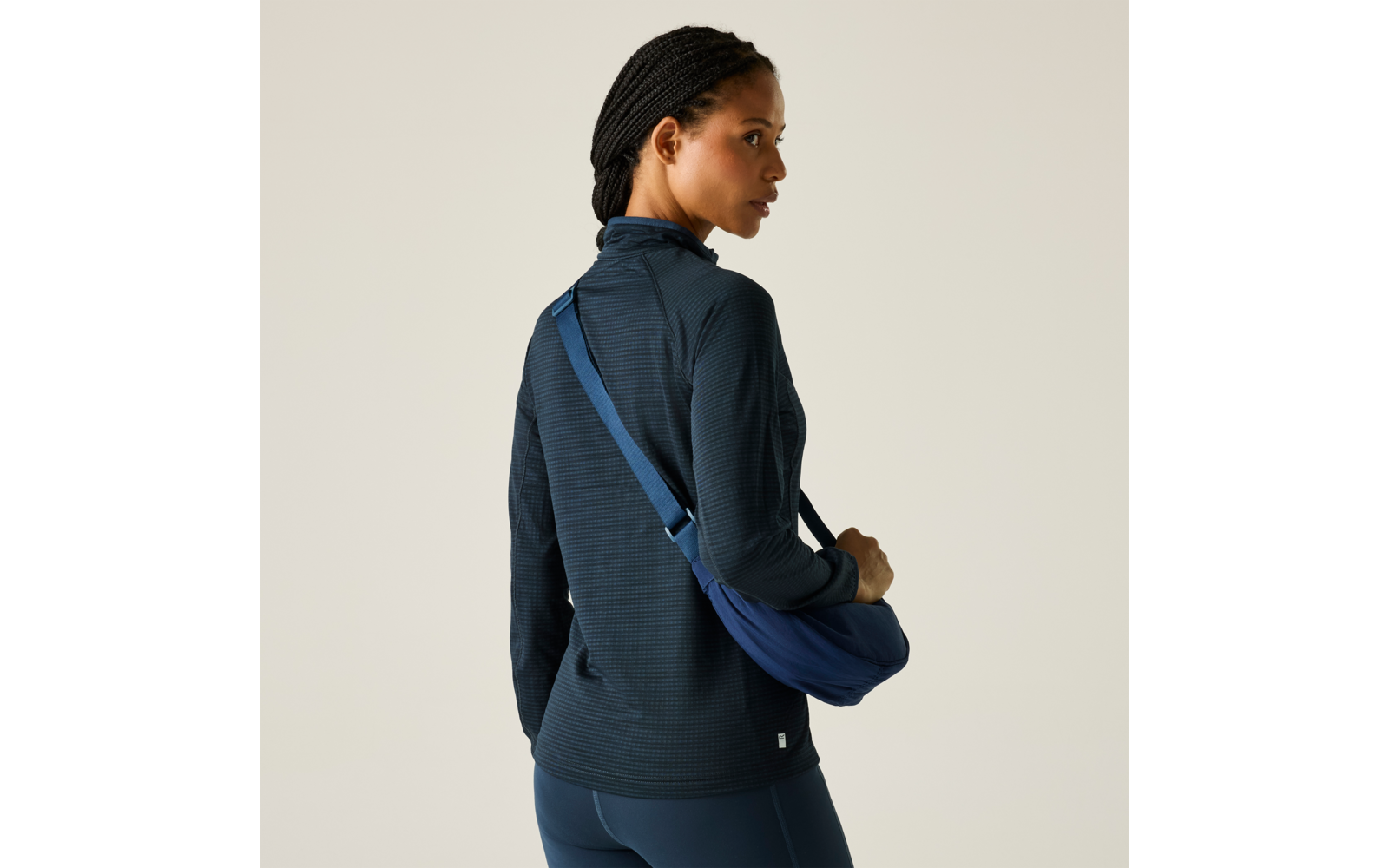 Femme de dos, légèrement tournée vers la gauche, portant un haut sombre à manches longues, des leggings bleu foncé et un sac bandoulière bleu en travers du dos, cheveux tressés, fond clair uni