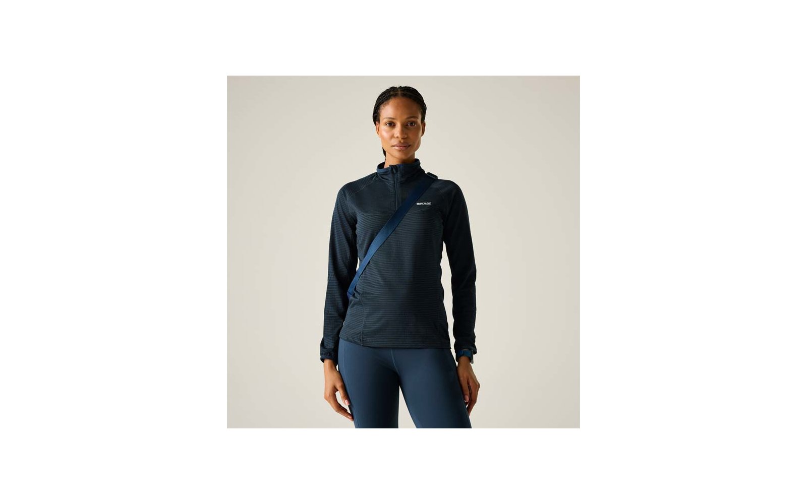 Personne debout de face, porte une veste zippée sombre à manches longues, leggings ajustés, bandoulière diagonale, cheveux nattés, fond clair