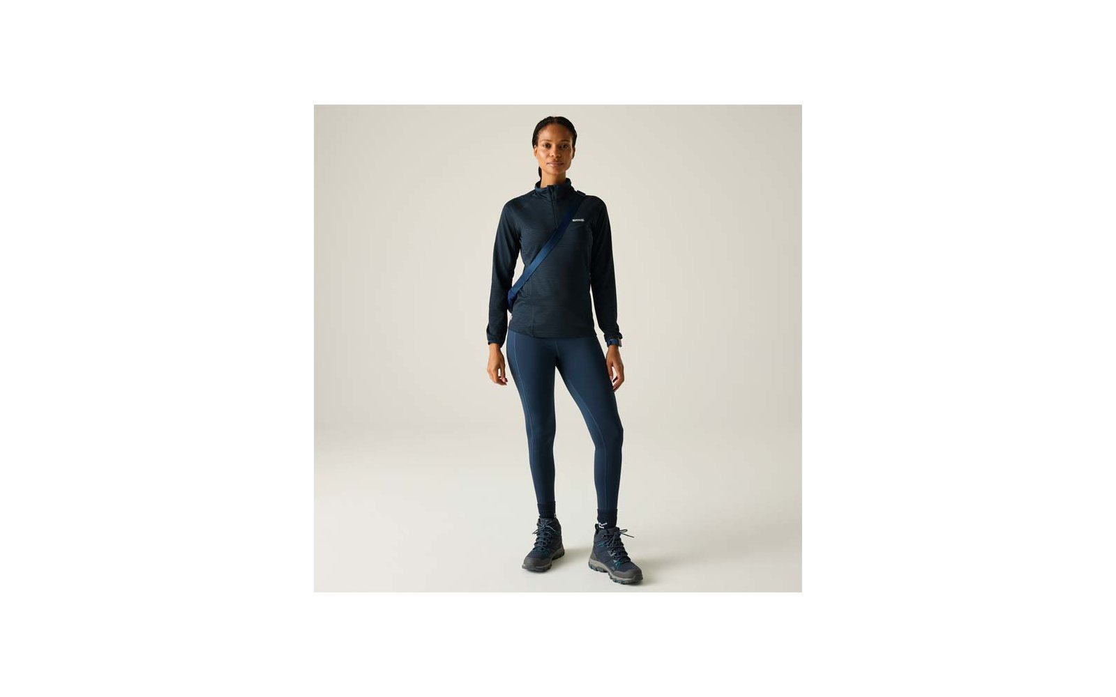 Personne debout de face sur fond clair, portant un haut de sport sombre à manches longues avec fermeture éclair, un legging sombre, une bandoulière en travers de la poitrine et des chaussures de randonnée