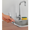 Hand hält ein graues und weißes kleines Gerät mit schmaler borstiger Spitze an der Kante eines Edelstahlspülbeckens; verchromter Wasserhahn, gelber Schwamm mit grünem Scheuerrand auf der Ablage, Holzarbeitsplatte, weiße Fliesen