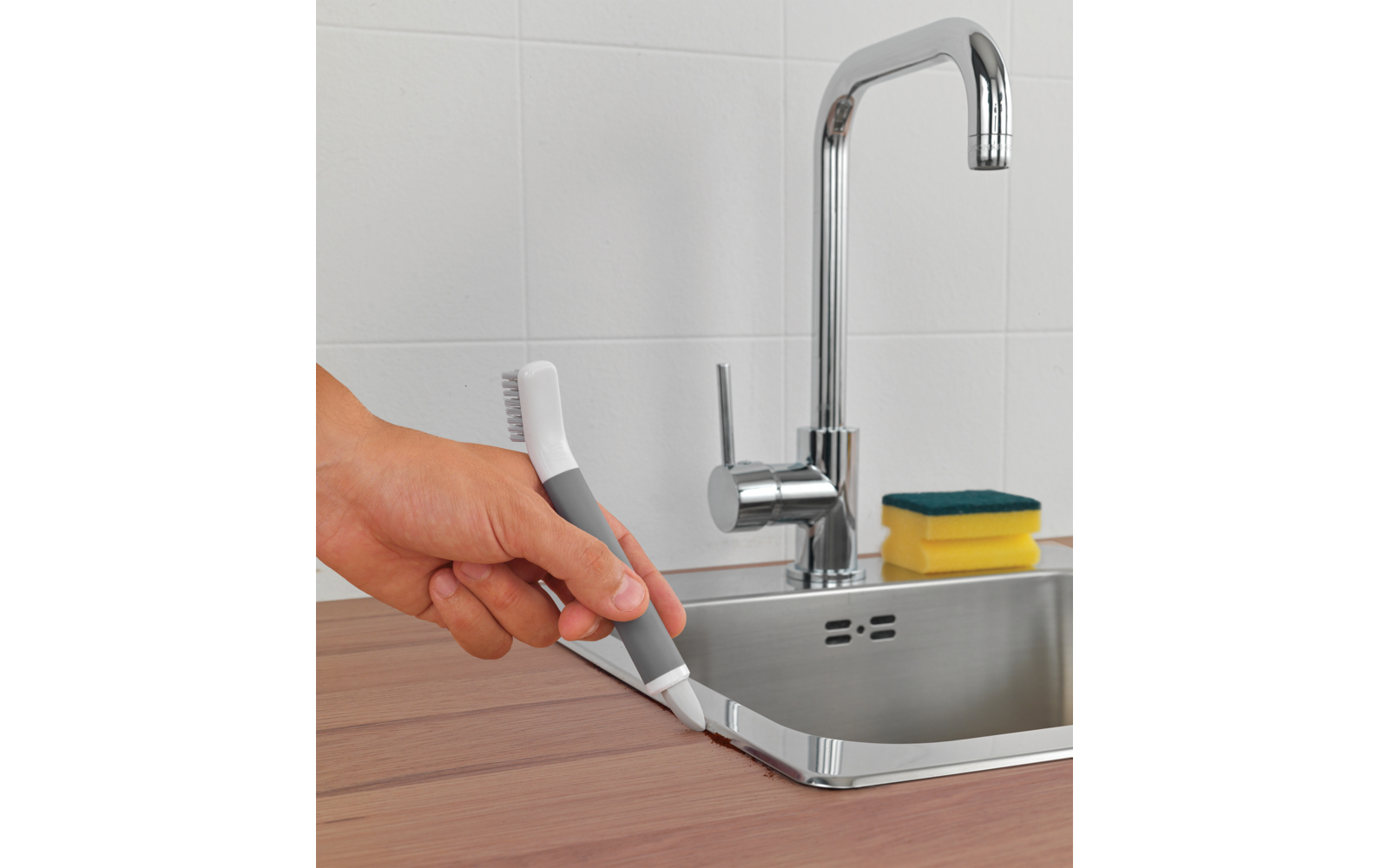 Hand hält ein graues und weißes kleines Gerät mit schmaler borstiger Spitze an der Kante eines Edelstahlspülbeckens; verchromter Wasserhahn, gelber Schwamm mit grünem Scheuerrand auf der Ablage, Holzarbeitsplatte, weiße Fliesen