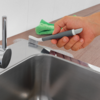Hand hält eine kleine graue Bürste mit weißen Borsten an der Kante einer Edelstahlspüle; grünes Mikrofasertuch liegt auf der Arbeitsplatte; Chromarmatur sichtbar