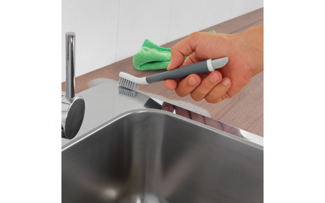 Hand hält eine kleine graue Bürste mit weißen Borsten an der Kante einer Edelstahlspüle; grünes Mikrofasertuch liegt auf der Arbeitsplatte; Chromarmatur sichtbar