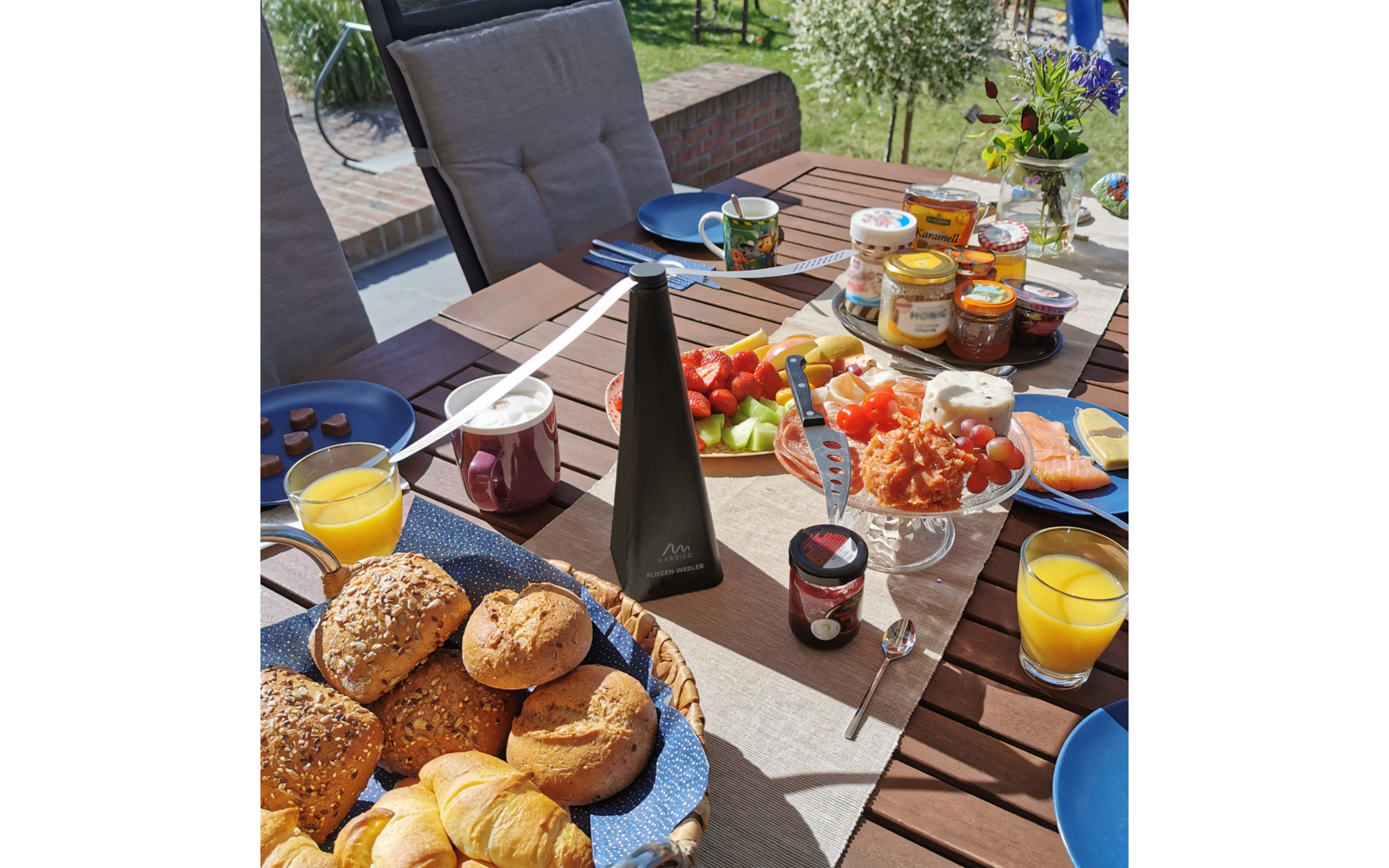 Ein gedeckter Gartentisch mit Brötchen, Gläsern mit Orangensaft, Marmeladengläsern und Schalen mit Erdbeeren