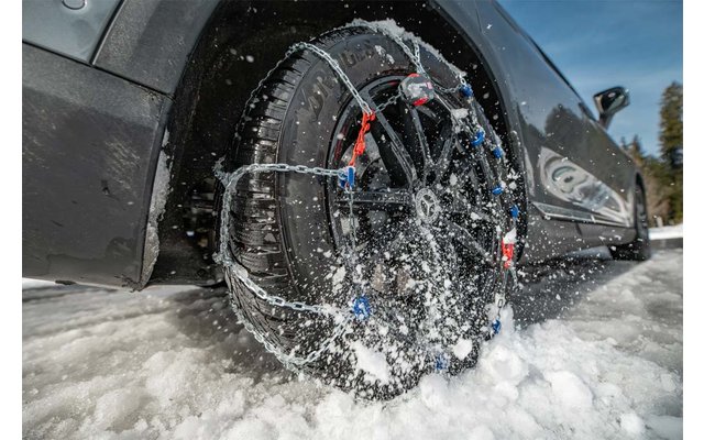 Primer plano de un neumático de coche con cadenas para la nieve, salpicaduras de agua, coche negro y nieve en la carretera