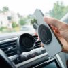 Hand hält ein Smartphone mit runder Befestigungsplatte und sichtbaren Kameralinsen vor einer schwarzen Autolüftungs-Handyhalterung am Armaturenbrett