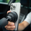 Hand hält silberweißes Smartphone mit drei Kameralinsen, befestigt an schwarzer Saugnapf-Halterung; unscharfer Autoinnenraum im Hintergrund