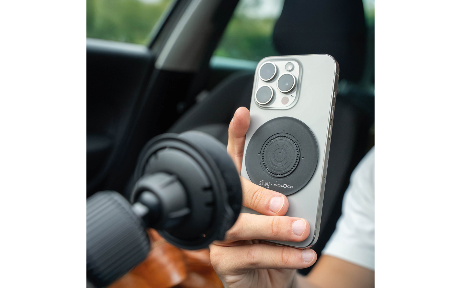 Hand hält ein silbernes Smartphone mit rundem schwarzem Pad auf der Rückseite, Autoinnenraum im Hintergrund