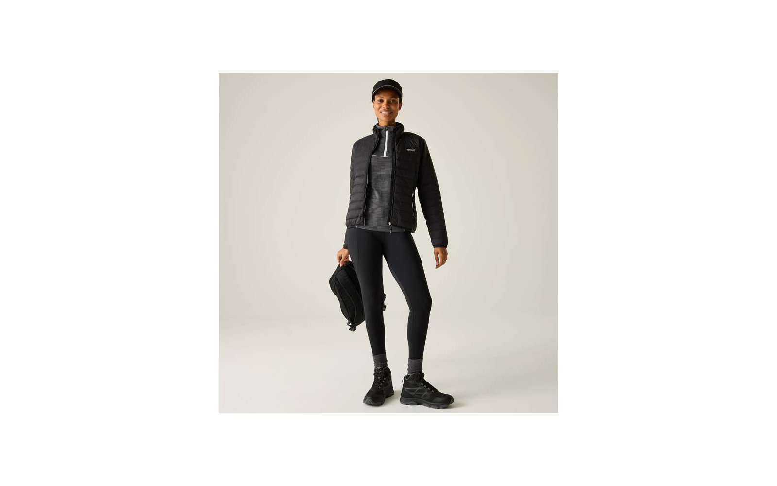 Person steht, trägt schwarze gesteppte Jacke mit Reißverschluss, grauen Pullover, schwarze Leggings, schwarze Wanderschuhe und schwarze Kappe, hält einen schwarzen Rucksack in der rechten Hand