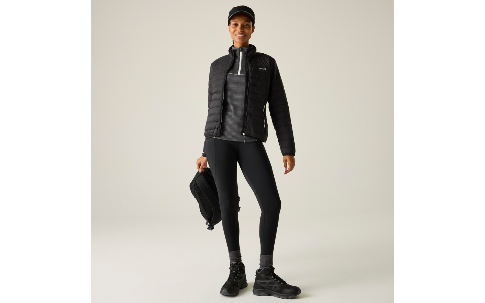 Person steht vor hellem Hintergrund, trägt schwarze Kappe, gesteppte schwarze Jacke, grauen Pullover mit Reißverschluss, schwarze Leggings und schwarze Wanderschuhe, hält einen schwarzen Rucksack in der linken Hand