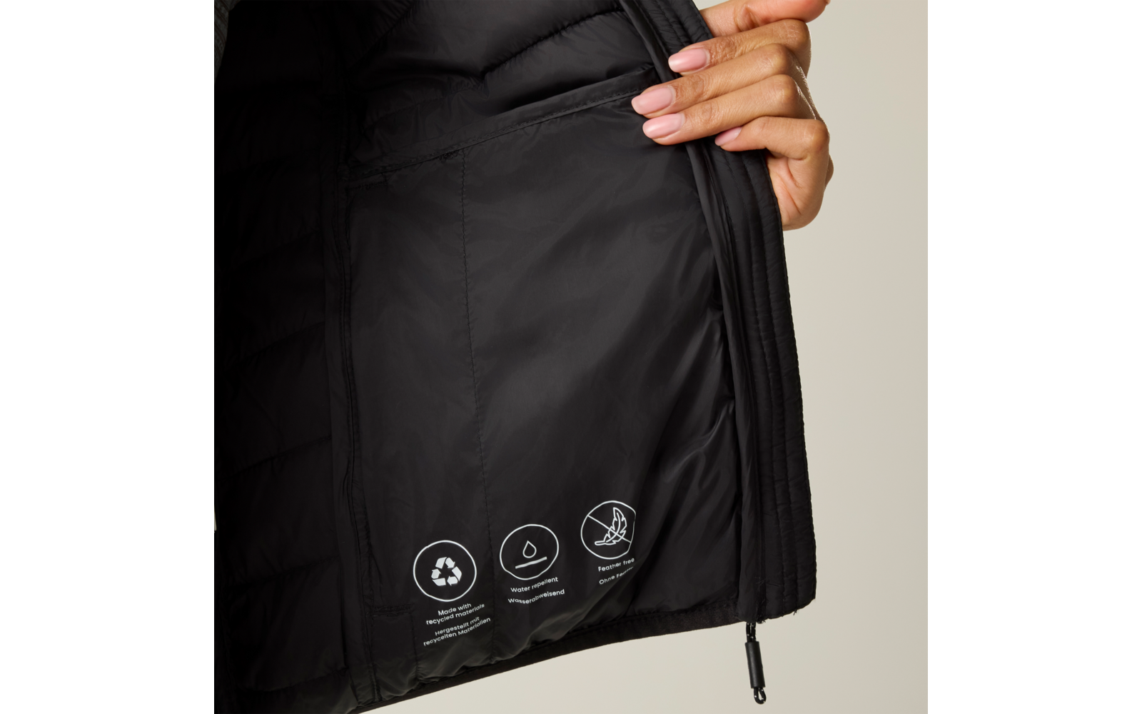 Hand hält die geöffnete Innenseite einer schwarzen gestepp­ten Jacke; innen drei weiße runde Symbole mit kleinem Text; sichtbarer schwarzer Reißverschluss mit Zug