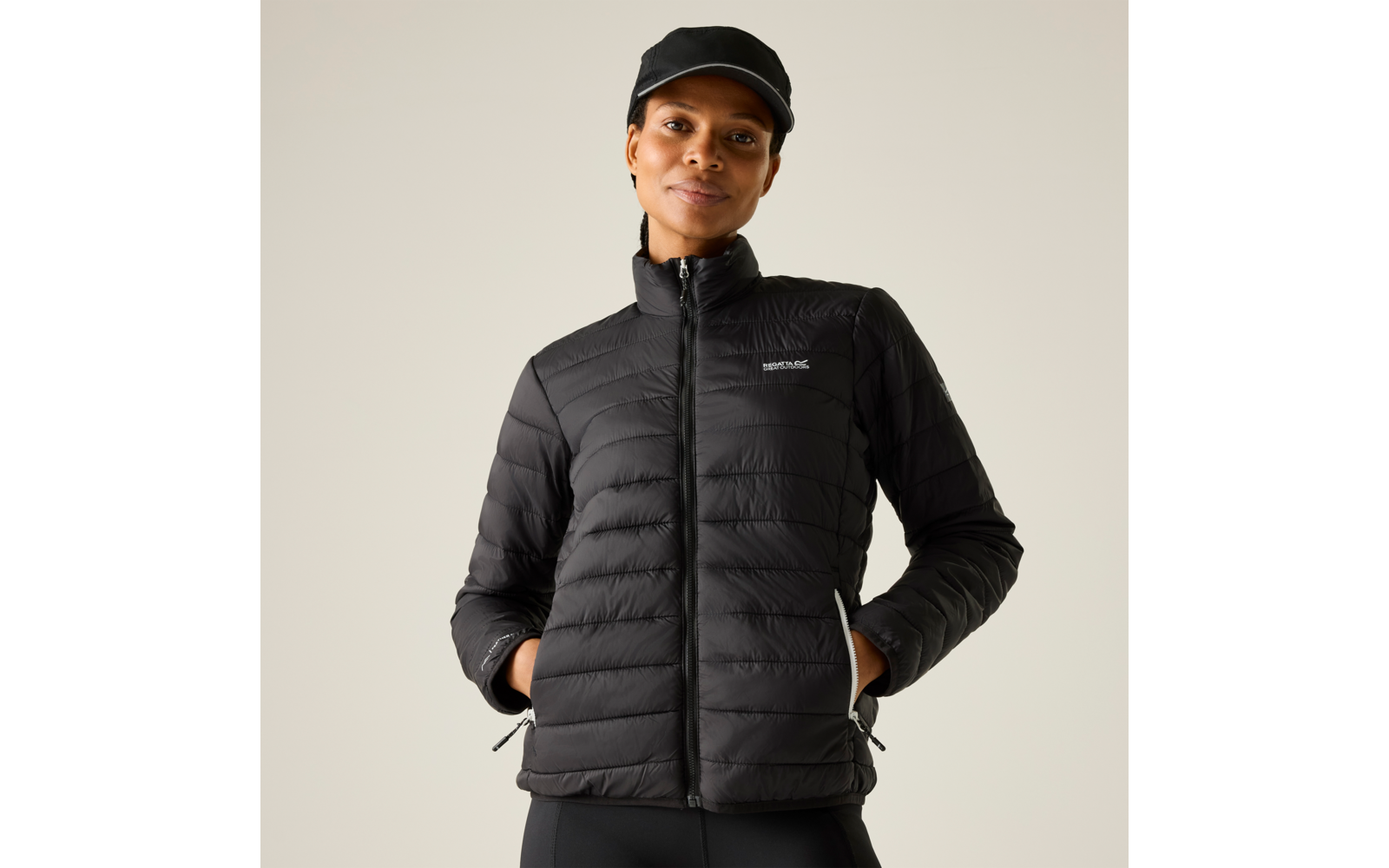 Person in schwarzer gesteppter Jacke und schwarzer Kappe, Hände in den Taschen, heller einfarbiger Hintergrund