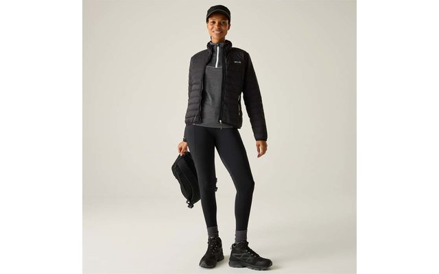 Person steht, trägt schwarze gesteppte Jacke mit Reißverschluss, grauen Pullover, schwarze Leggings, schwarze Wanderschuhe und schwarze Kappe, hält einen schwarzen Rucksack in der rechten Hand