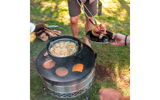 Hand mit Zange legt einen belegten Burger auf einen Teller neben einem runden Außengrill; auf dem Grill eine Pfanne mit bratenden Kartoffelspalten, zwei Brötchenhälften und eine Käsescheibe; Beine in Shorts und Holzbündel im Gras