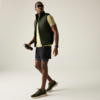 Persoon in zijaanzicht, draagt zonnebril, doorgestikte mouwloze bodywarmer, lichtgroen T‑shirt met korte mouwen, zwarte shorts en groene sneakers; houdt een zwarte drinkfles in de rechterhand; draagt horloge; effen lichte achtergrond