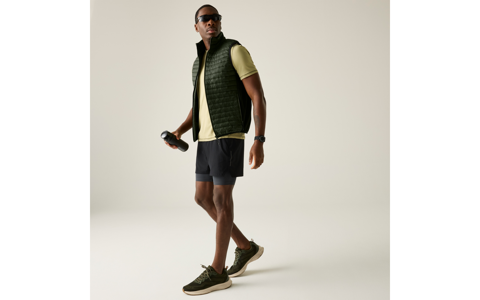 Persoon in zijaanzicht, draagt zonnebril, doorgestikte mouwloze bodywarmer, lichtgroen T‑shirt met korte mouwen, zwarte shorts en groene sneakers; houdt een zwarte drinkfles in de rechterhand; draagt horloge; effen lichte achtergrond
