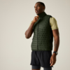 Persoon staat, draagt donkergroene doorgestikte bodywarmer over lichtgroen T‑shirt met korte mouwen, zwarte korte broek en polshorloge; één hand houdt de rits bij de kraag; effen lichte achtergrond