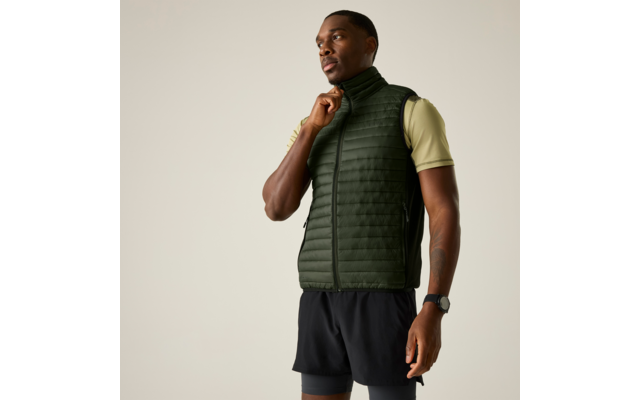 Persoon staat, draagt donkergroene doorgestikte bodywarmer over lichtgroen T‑shirt met korte mouwen, zwarte korte broek en polshorloge; één hand houdt de rits bij de kraag; effen lichte achtergrond