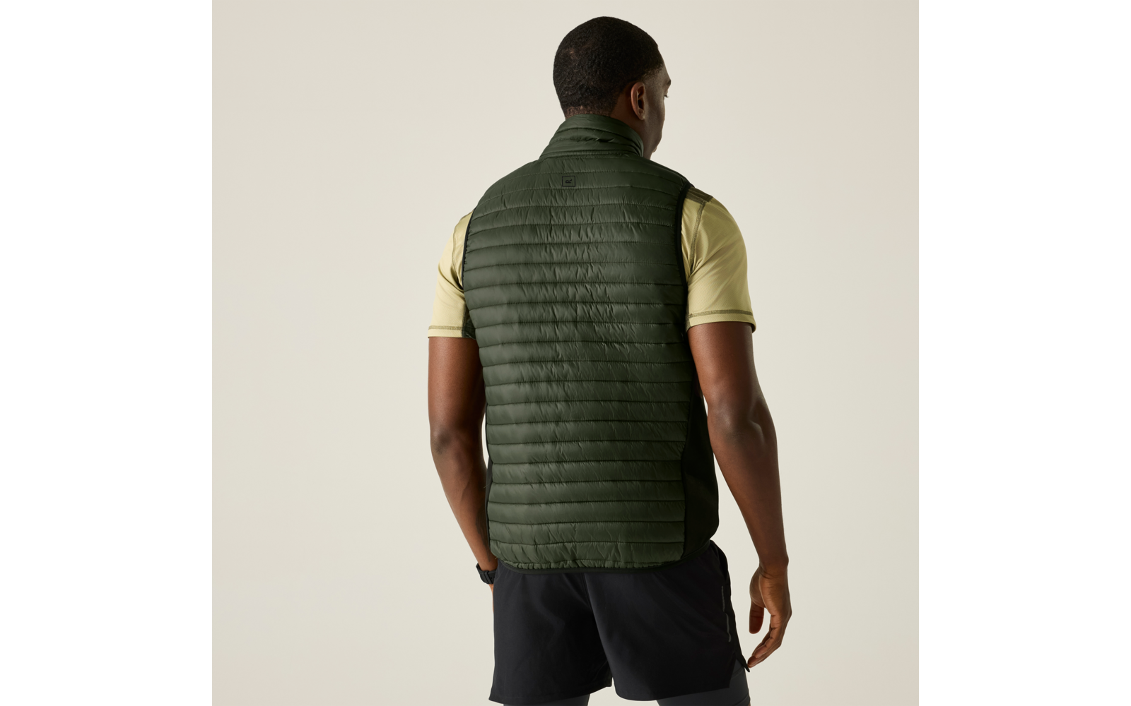 Man van achteren, draagt een donkergroene doorgestikte bodywarmer over een lichtgeel T‑shirt met korte mouwen en donkere korte broek op een effen lichte achtergrond