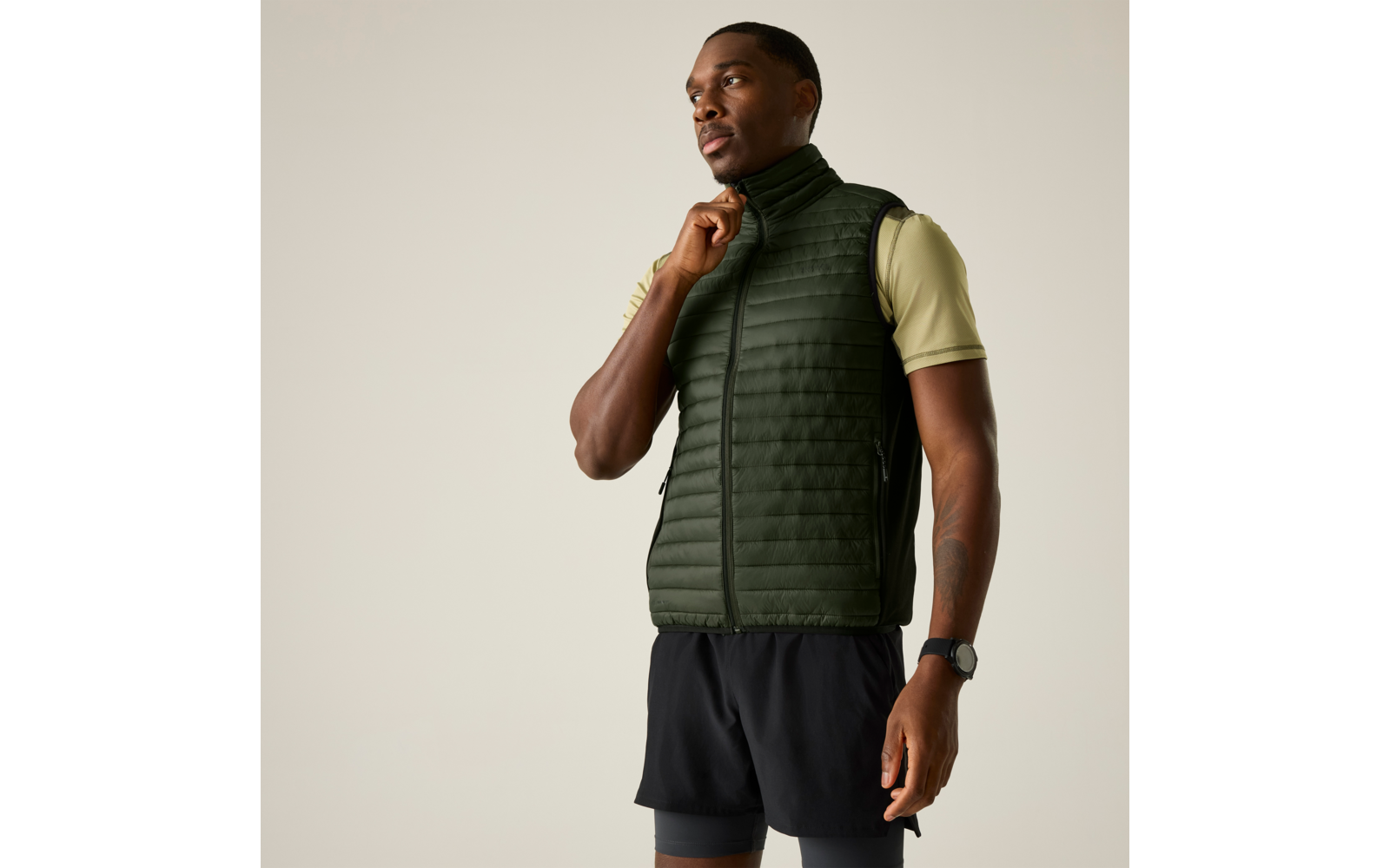 Persoon staat, draagt donkergroene doorgestikte bodywarmer over lichtgroen T‑shirt met korte mouwen, zwarte korte broek en polshorloge; één hand houdt de rits bij de kraag; effen lichte achtergrond